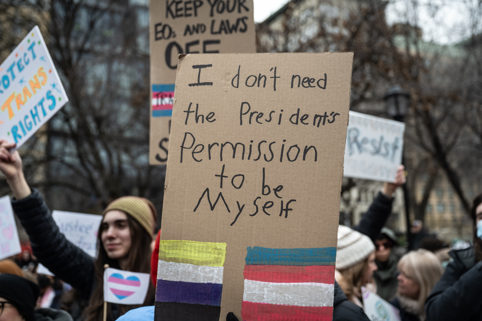 A protester holds up a sign that reads "I don't need the president's permission to be myself" with a painting of the trans and nonbinary flags.