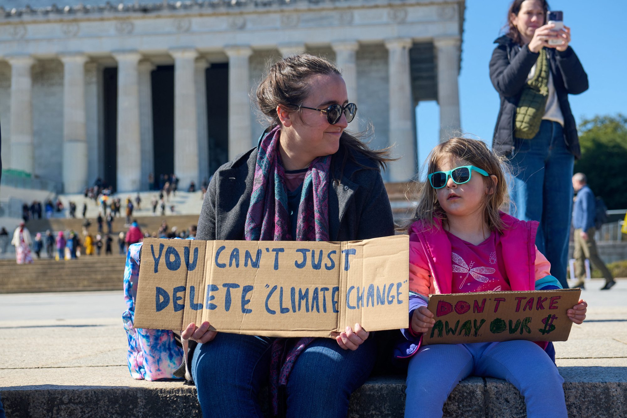 A woman and child sit on the steps outside the Lincoln Memorial. One holds a sign that reads "You can't just delete climate change." The other holds one reading "Don't take away our $".