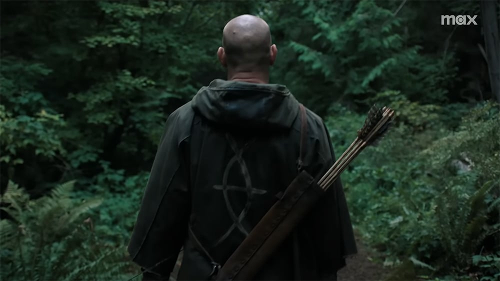 A man in a hooded robe carrying a quiver of arrows is seen from behind walking through the woods.