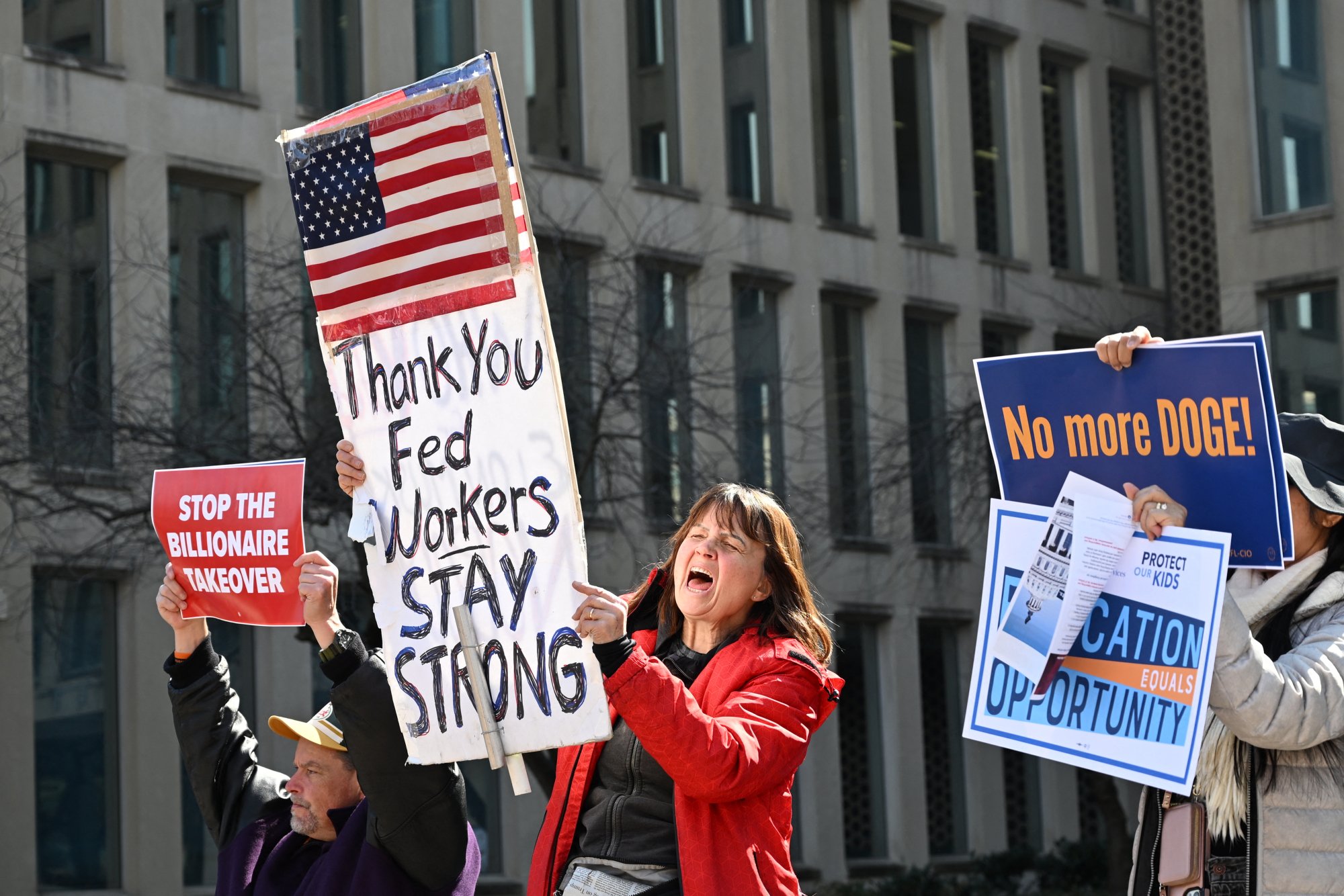 A small group of protesters hold signs outside the Office of Personnel Management in D.C. One reads "Thank you fed workers stay strong." Another reads "No more DOGE!"