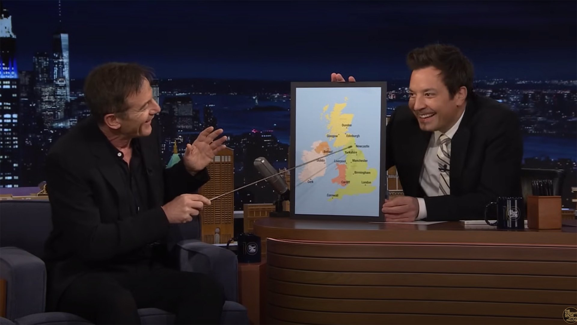 Two men sit on a talk show with a map of the UK between them. The man on the left uses a pointer to point at the map.