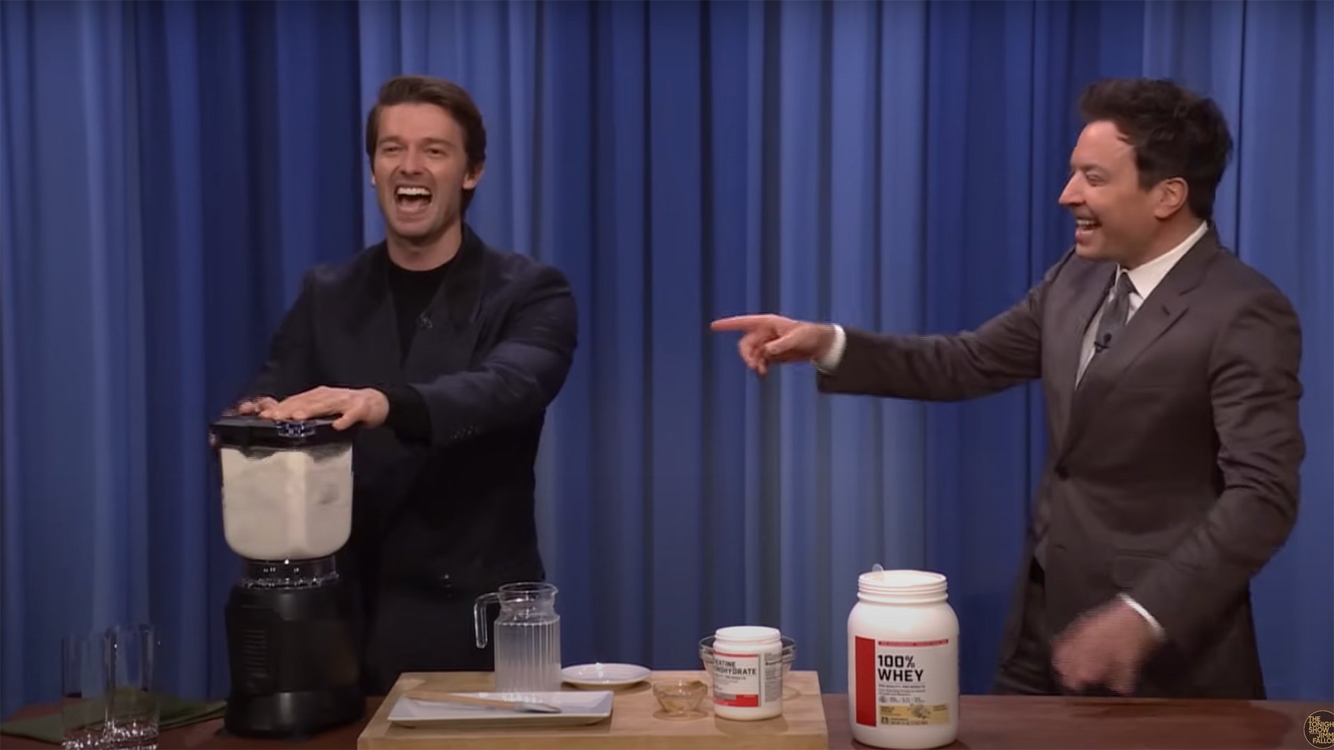 Two men stand on a stage making a protein shake in a blender, laughing.